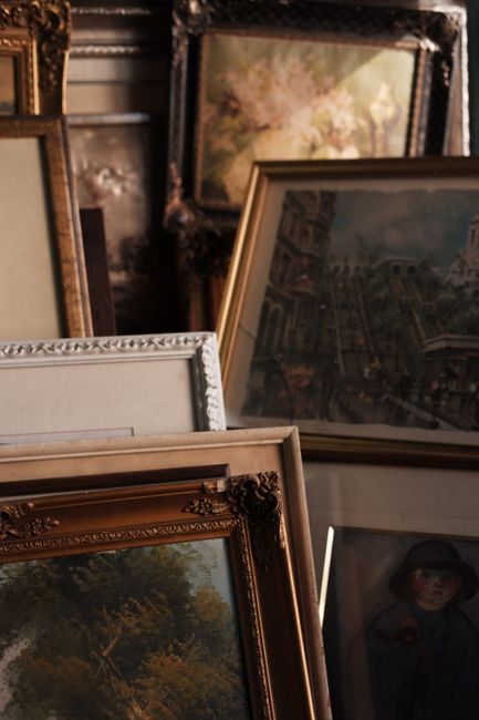 A collection of framed artwork and paintings in various sizes and ornate frames, leaning against each other in a storage or preparation area. The images include landscapes, cityscapes, and portraits,